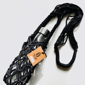 May include: Black macrame water bottle holder with a clear glass bottle and silver lid. The holder has a long strap for carrying. A brown tag with the text "Organically Pineapple" is attached.