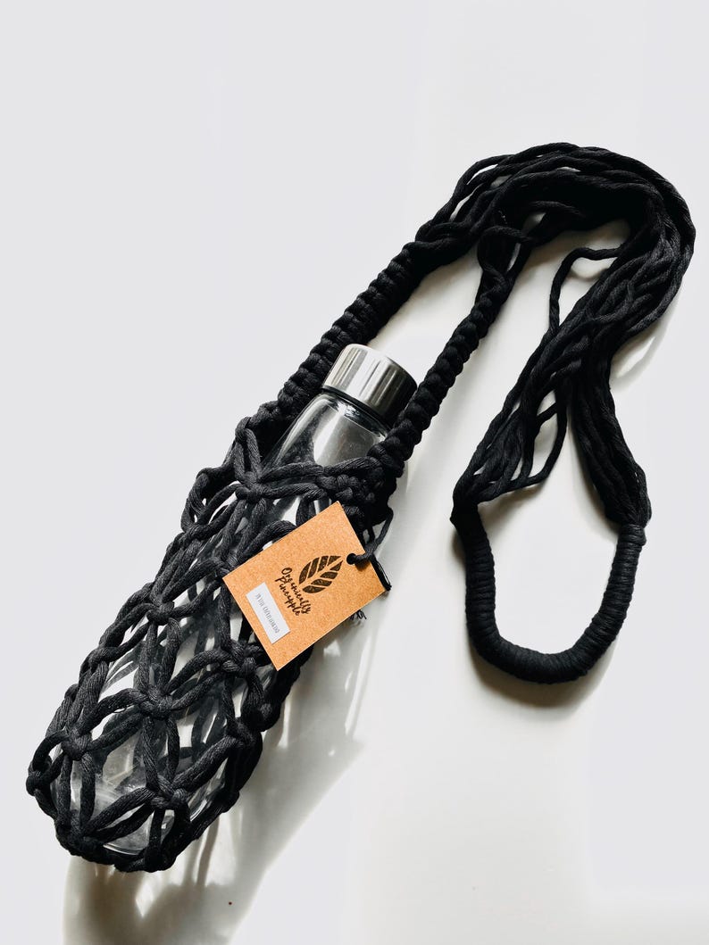 May include: Black macrame water bottle holder with a clear glass bottle and silver lid. The holder has a long strap for carrying. A brown tag with the text "Organically Pineapple" is attached.