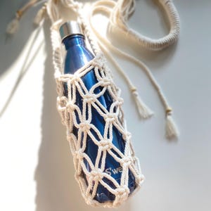 May include: A blue water bottle in a cream-colored macrame holder with a long strap. The bottle has a silver lid and the macrame has a woven net design. The word "S'well" is visible on the bottle.