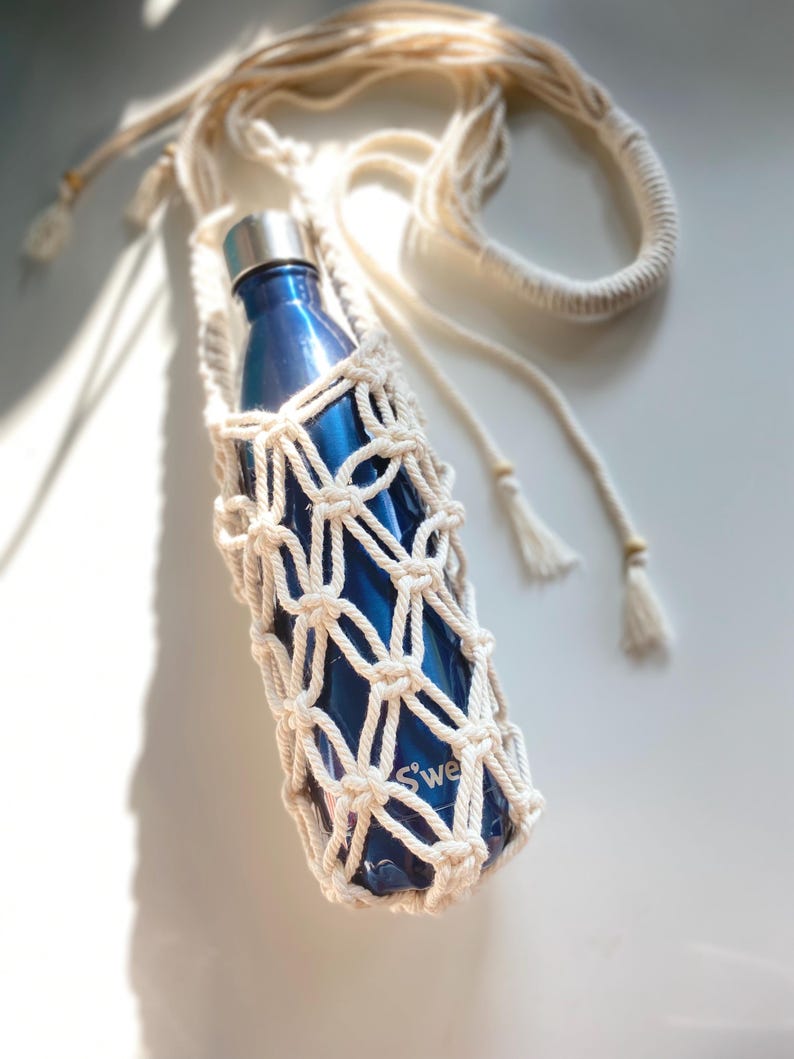 May include: A blue water bottle in a cream-colored macrame holder with a long strap. The bottle has a silver lid and the macrame has a woven net design. The word "S'well" is visible on the bottle.