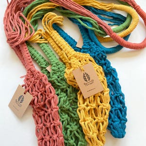 May include: A collection of colorful macrame plant hangers in shades of coral, green, yellow, and teal. Each hanger has a woven net design and a long, looped handle. Small tags with a leaf logo are attached.