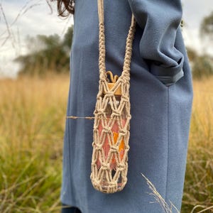 May include: A clear glass water bottle with orange and pink contents is held in a beige macrame carrier with a long strap. The bottle has a wooden cap. The carrier is hanging against a blue coat in a field of tall grass.