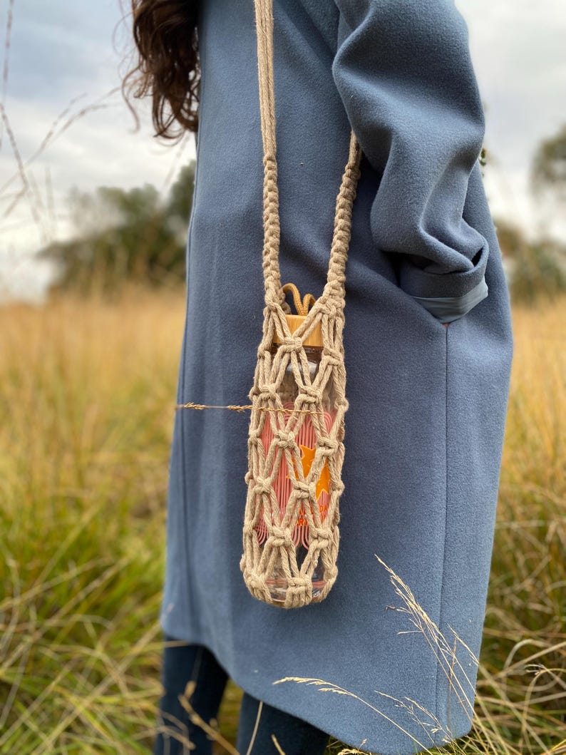 May include: A clear glass water bottle with orange and pink contents is held in a beige macrame carrier with a long strap. The bottle has a wooden cap. The carrier is hanging against a blue coat in a field of tall grass.