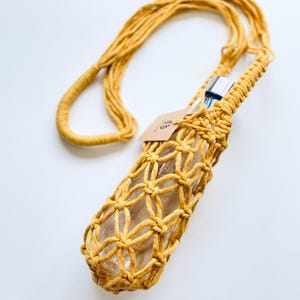 May include: Mustard yellow macrame water bottle holder with a long strap. The holder is designed to fit a clear glass bottle. A small tag is attached to the holder. The background is white.