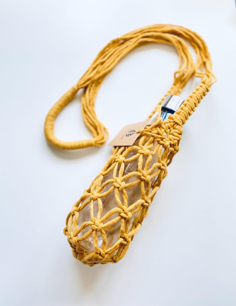May include: Mustard yellow macrame water bottle holder with a long strap. The holder is designed to fit a clear glass bottle. A small tag is attached to the holder. The background is white.