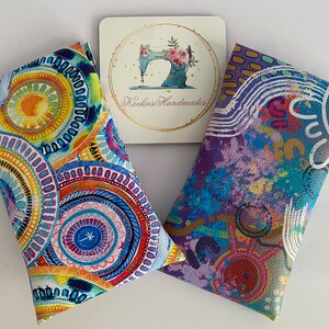 May include: Two colorful eyeglass cases with abstract designs. One case features a circular pattern in shades of blue, yellow, and orange. The other case has a purple and blue abstract design. A small card with a sewing machine logo is between them.