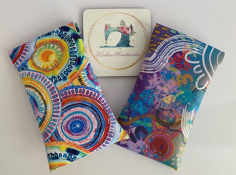 May include: Two colorful eyeglass cases with abstract designs. One case features a circular pattern in shades of blue, yellow, and orange. The other case has a purple and blue abstract design. A small card with a sewing machine logo is between them.