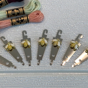 May include: Silver-coloured thread organisers, each with a gold-coloured bee decoration. Several spools of embroidery floss in purple, green, and peach are also visible, suggesting a crafting theme.