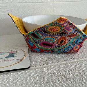 May include: A white ceramic bowl is nestled in a patterned fabric bowl cosy. The cosy displays a colourful design of blue, red, orange, and purple dots on a black backdrop. A coaster with a sewing machine design is also present.