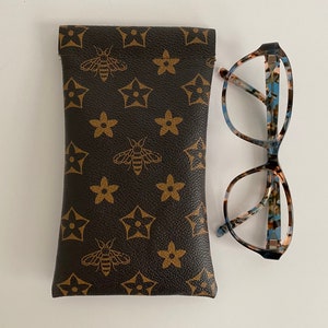 May include: A brown glasses case featuring a bee and star pattern, alongside a pair of patterned spectacles. The case is rectangular and has a leather-like texture. The glasses have a tortoiseshell design.