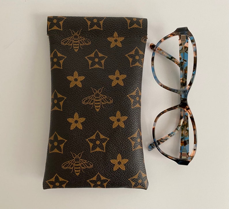 May include: A brown glasses case featuring a bee and star pattern, alongside a pair of patterned spectacles. The case is rectangular and has a leather-like texture. The glasses have a tortoiseshell design.
