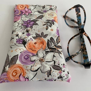 May include: A floral eyeglasses case with a gray background and orange, purple, and white flowers. The case is next to a pair of eyeglasses with a patterned frame.