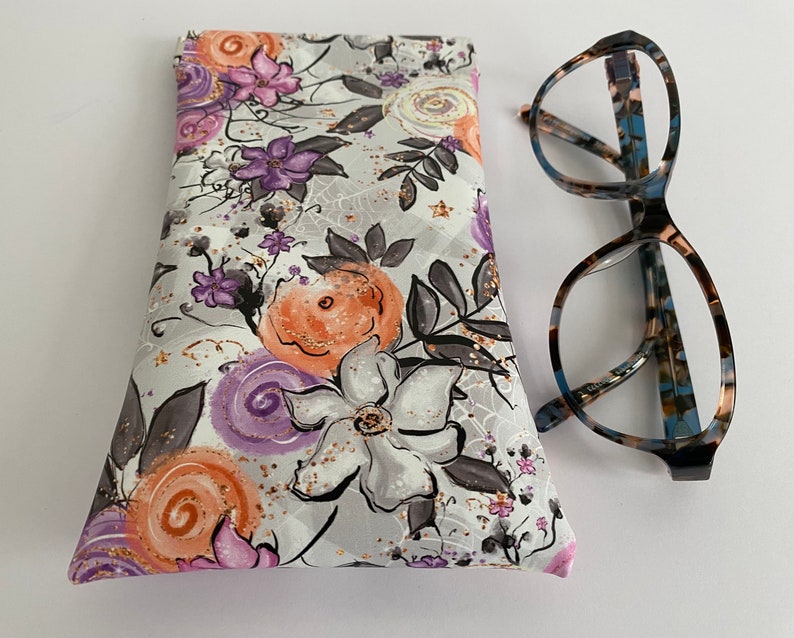 May include: A floral eyeglasses case with a gray background and orange, purple, and white flowers. The case is next to a pair of eyeglasses with a patterned frame.