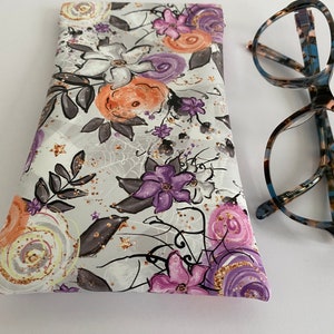 May include: A floral eyeglasses case with a gray background and a pattern of purple, orange, and white flowers. The case is next to a pair of eyeglasses with a tortoiseshell pattern frame.