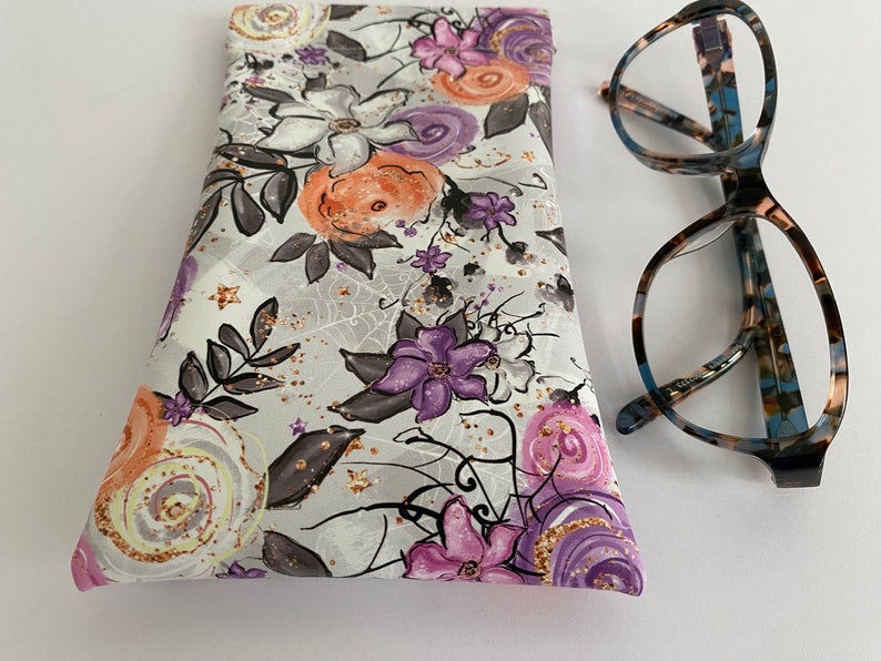 May include: A floral eyeglasses case with a gray background and a pattern of purple, orange, and white flowers. The case is next to a pair of eyeglasses with a tortoiseshell pattern frame.