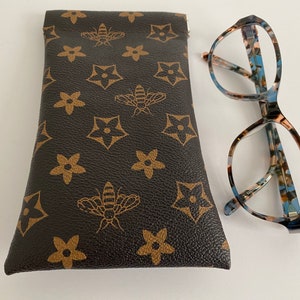 May include: Eyeglasses with a patterned frame in blue, brown, and cream, beside a brown glasses case. The case features a repeating pattern of bees and star-shaped flowers.