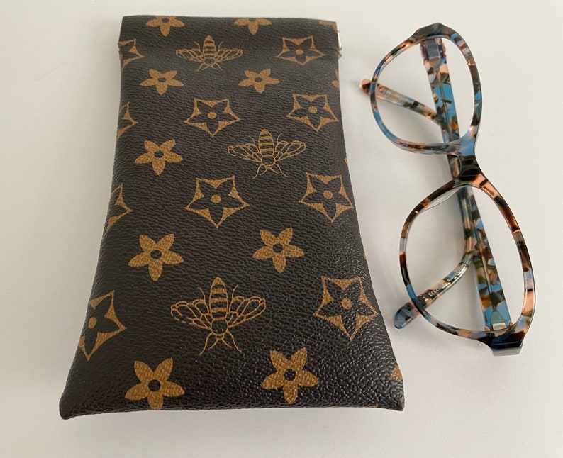 May include: Eyeglasses with a patterned frame in blue, brown, and cream, beside a brown glasses case. The case features a repeating pattern of bees and star-shaped flowers.
