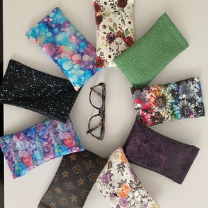 May include: A selection of spectacle cases in diverse patterns and colours, arranged in a circular pattern around a pair of spectacles. The cases feature designs such as floral prints, abstract patterns, and textured surfaces.