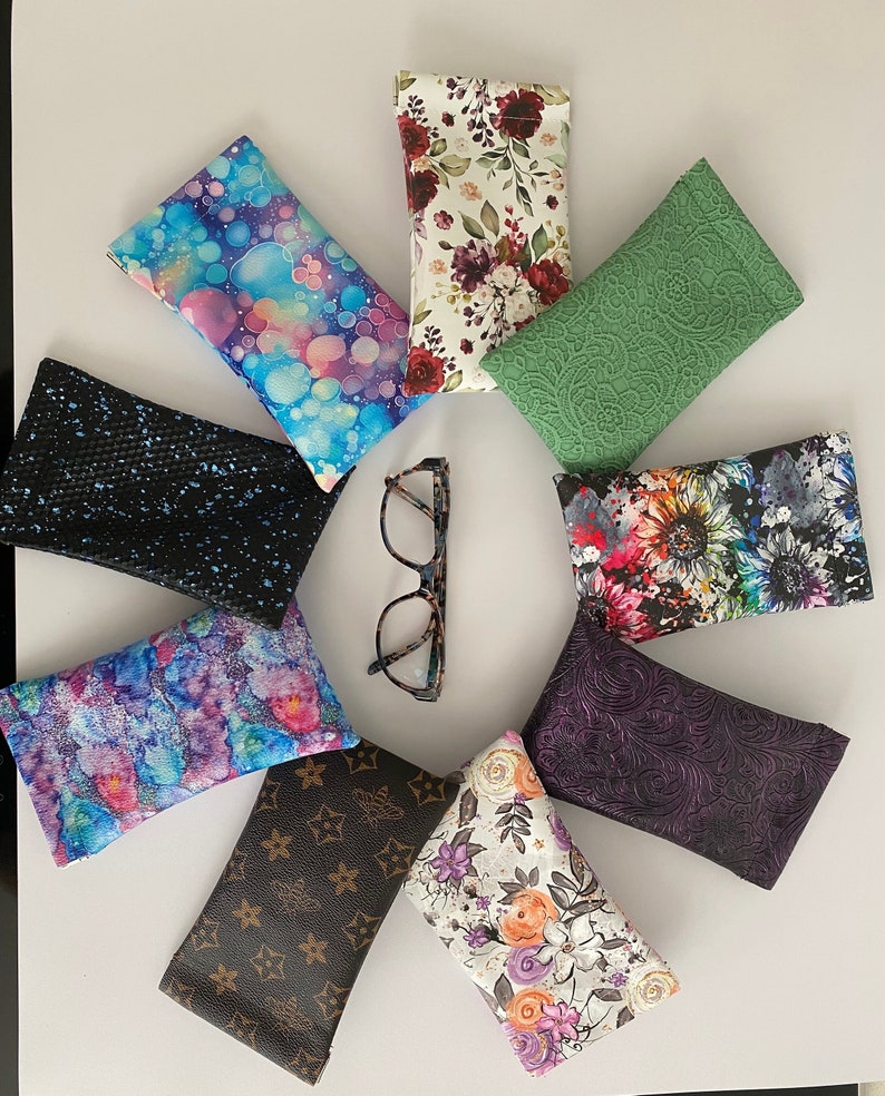 May include: A selection of spectacle cases in diverse patterns and colours, arranged in a circular pattern around a pair of spectacles. The cases feature designs such as floral prints, abstract patterns, and textured surfaces.