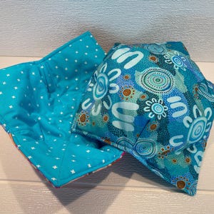 May include: Two fabric bowl cozies. One is open, showing a turquoise interior with white crosses. The other is folded, displaying a blue pattern with white, brown, and turquoise abstract designs. These cozies are designed to keep bowls warm.