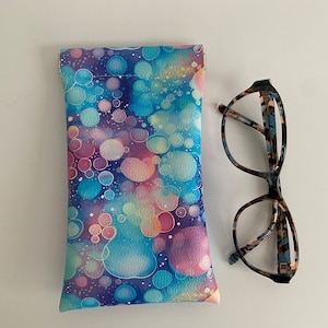 May include: A vibrant glasses case featuring a pattern of overlapping circles in blue, pink, and yellow hues. Beside it, a pair of spectacles with a tortoiseshell frame.