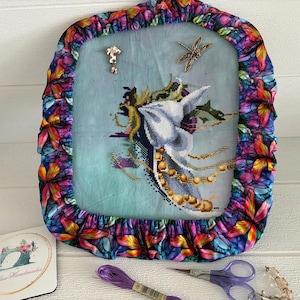 May include: Cross-stitch project depicting a mythical creature with a white head and gold detailing, framed by a vibrant floral fabric. Accessories include scissors, thread, and a decorative pin. The project is displayed on a white surface.
