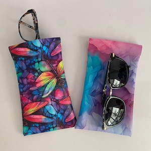 May include: Two eyeglass cases. One case has a colourful dragonfly design with a stained glass effect. The other case has a watercolour-style design in shades of pink, purple, and blue. Sunglasses are in the second case, and eyeglasses are in the first.