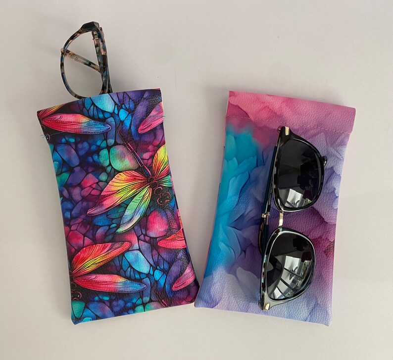 May include: Two eyeglass cases. One case has a colourful dragonfly design with a stained glass effect. The other case has a watercolour-style design in shades of pink, purple, and blue. Sunglasses are in the second case, and eyeglasses are in the first.