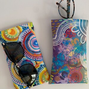 May include: Two eyeglass cases. One case is patterned with a colorful circular design in shades of blue, yellow, and orange. The other case has a purple, blue, and red abstract design. Sunglasses and eyeglasses are in the cases.