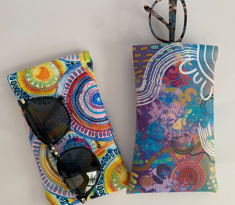 May include: Two eyeglass cases. One case is patterned with a colorful circular design in shades of blue, yellow, and orange. The other case has a purple, blue, and red abstract design. Sunglasses and eyeglasses are in the cases.