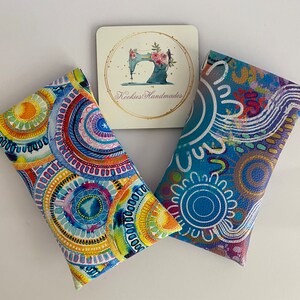 May include: Two rectangular fabric cases with colorful, abstract designs. One features concentric circles in blue, yellow, and red. The other has a blue background with white and purple patterns. A small card with a sewing machine logo and the text "Kookies Handmades" is also present.