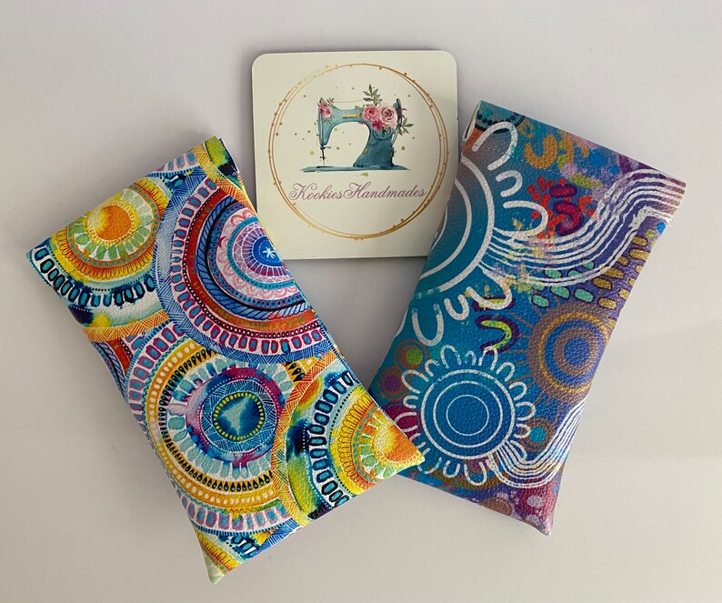 May include: Two rectangular fabric cases with colorful, abstract designs. One features concentric circles in blue, yellow, and red. The other has a blue background with white and purple patterns. A small card with a sewing machine logo and the text "Kookies Handmades" is also present.