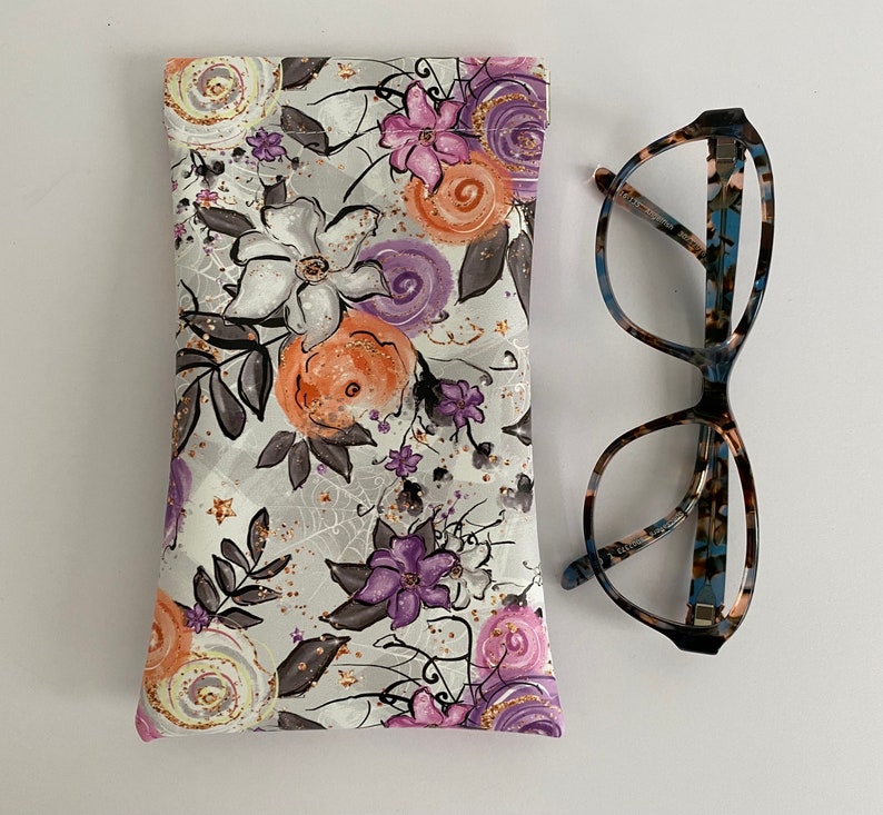 May include: A floral print glasses case with a matching pair of eyeglasses. The case features a pattern of white, orange, and purple flowers with gray leaves on a light gray background. The eyeglasses have a tortoiseshell pattern.