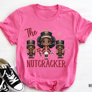 May include: Pink t-shirt with a graphic of three cartoon nutcrackers. The center nutcracker is a girl with brown skin and black hair. The text "The Nutcracker" is printed below the graphic.