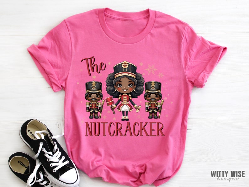 May include: Pink t-shirt with a graphic of three cartoon nutcrackers. The center nutcracker is a girl with brown skin and black hair. The text "The Nutcracker" is printed below the graphic.