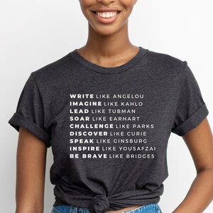 May include: A dark grey t-shirt with white text that reads "WRITE LIKE ANGELOU IMAGINE LIKE KAHLO LEAD LIKE TUBMAN SOAR LIKE EARHART CHALLENGE LIKE PARKS DISCOVER LIKE CURIE SPEAK LIKE GINSBURG INSPIRE LIKE YOUSAFZAI BE BRAVE LIKE BRIDGES"