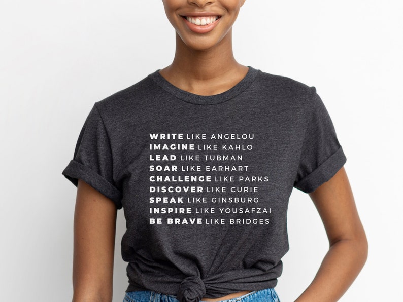 May include: A dark grey t-shirt with white text that reads "WRITE LIKE ANGELOU IMAGINE LIKE KAHLO LEAD LIKE TUBMAN SOAR LIKE EARHART CHALLENGE LIKE PARKS DISCOVER LIKE CURIE SPEAK LIKE GINSBURG INSPIRE LIKE YOUSAFZAI BE BRAVE LIKE BRIDGES"
