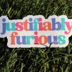 May include: A white sticker with the text "justifiably furious" in rainbow colours with glitter accents.