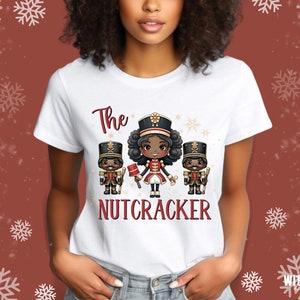 May include: White t-shirt with a graphic of three cartoon nutcrackers. The center nutcracker is a girl with brown skin and brown hair. The text "The Nutcracker" is printed below the graphic.