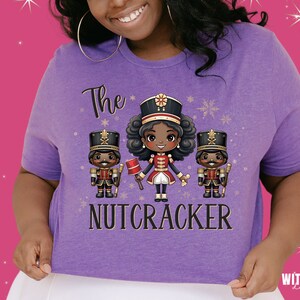 May include: Purple t-shirt with a graphic of three cartoon nutcrackers. The center nutcracker is a girl with brown hair and a red and gold uniform. The text "The Nutcracker" is printed below the graphic.