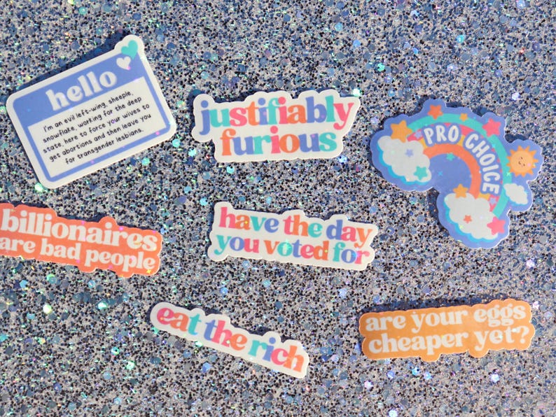 May include: A collection of colourful stickers with various messages, including "hello", "justifiably furious", "billionaires are bad people", "have the day you voted for", "eat the rich", "pro choice", and "are your eggs cheaper yet?"