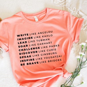 May include: A coral pink t-shirt with black text that reads "Write like Angelou, Imagine like Kahlo, Lead like Tubman, Soar like Earhart, Challenge like Parks, Discover like Curie, Speak like Ginsburg, Inspire like Yousafzai, Be Brave like Bridges."
