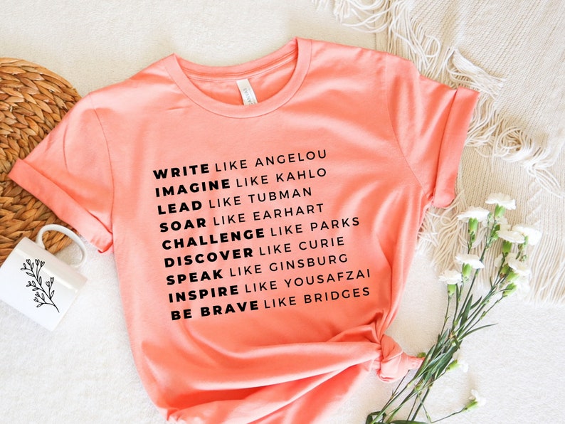 May include: A coral pink t-shirt with black text that reads "Write like Angelou, Imagine like Kahlo, Lead like Tubman, Soar like Earhart, Challenge like Parks, Discover like Curie, Speak like Ginsburg, Inspire like Yousafzai, Be Brave like Bridges."
