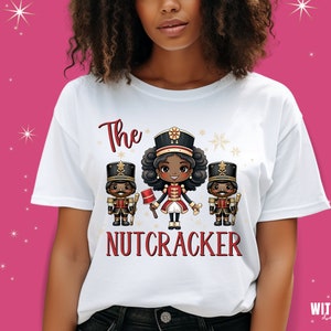 May include: White t-shirt with a graphic of three cartoon nutcrackers. The center nutcracker is a girl with brown skin and curly hair. The text "The Nutcracker" is printed below the graphic.