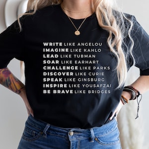 May include: Black t-shirt with white text that reads "Write like Angelou, Imagine like Kahlo, Lead like Tubman, Soar like Earhart, Challenge like Parks, Discover like Curie, Speak like Ginsburg, Inspire like Yousafzai, Be Brave like Bridges."