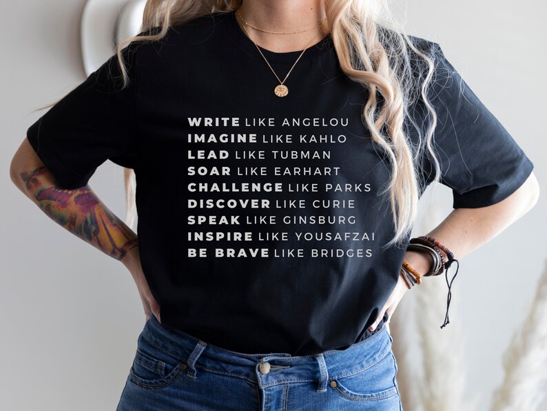 May include: Black t-shirt with white text that reads "Write like Angelou, Imagine like Kahlo, Lead like Tubman, Soar like Earhart, Challenge like Parks, Discover like Curie, Speak like Ginsburg, Inspire like Yousafzai, Be Brave like Bridges."