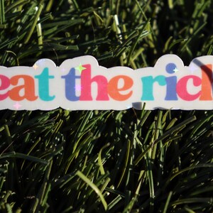 May include: A white sticker with the text "eat the rich" in rainbow colours. The sticker has a glossy finish and is on a green background.