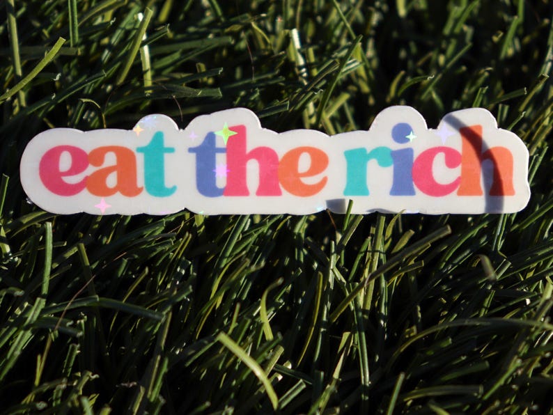 May include: A white sticker with the text "eat the rich" in rainbow colours. The sticker has a glossy finish and is on a green background.