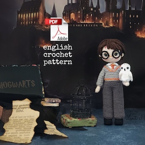 Könnte beinhalten: Häkelanleitung für eine Harry Potter Amigurumi-Puppe. Die Puppe trägt einen grauen Pullover und eine Hose mit einem rot-goldenen Schal. Die Puppe hält eine weiße Eule. Die Anleitung ist im PDF-Format verfügbar.