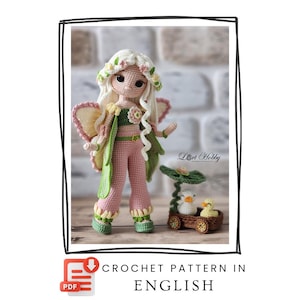 May include: A crochet doll with long white hair, a floral crown, pink pants, and green accents, featuring wings and a green top. Beside the doll is a small brown cart containing two white and yellow ducklings under a green leaf-shaped umbrella.